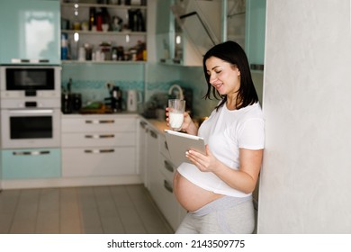 Pregnant Middle Aged Woman Using Digital Tablet While Having Milk In The Kitchen At Home. Expectant Mother Morning Routine, Breakfast, Healthy Lifestyle 