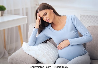 Pregnant Millennial Girl Having Headache Or Migraine Sitting On Sofa At Home. Pregnancy And Health Concept