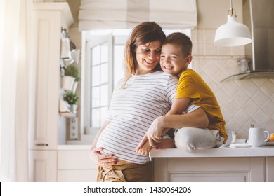 Pregnant Mom With Kid Playing Together And Laughing In The Kitchen. Mother With Son Sharing Good Emotions While Having Breakfast At Home.