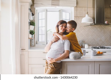 Mãe grávida com filho brincando juntos e se abraçando na cozinha. Mãe com filho compartilhando boas emoções enquanto toma café da manhã em casa.