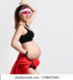 Pregnant mom in pink sun visor, black sport shorts and top. She is tying red hoody on her tummy, posing isolated on white background. Family and maternity concepts. Close up, copy space - Shutterstock ID 1708473463