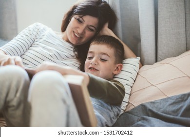 Pregnant Mom Reading Book And Talking With Her Child In The Bedroom. Mother With Son Sharing Good Emotions While Relaxing At Home.