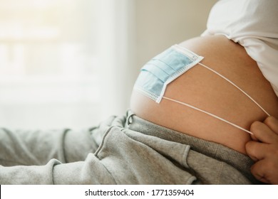 Pregnant Mother Asia Use Face Mask Covering On Her Abdomen For Protecting Fetus Or Baby Coronavirus, Pollution, Flu Or Pandemic. Pregnancy Woman Quarantine At Home. Afraid Spread Coronavirus Covid 19