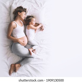 Pregnant Mother And Daughter Resting In Bed At Home. Young Woman With Her First Child During Second Pregnancy. Motherhood And Parenting Concept. Toddler Girl And Mom. Happy Family Expecting For Baby.