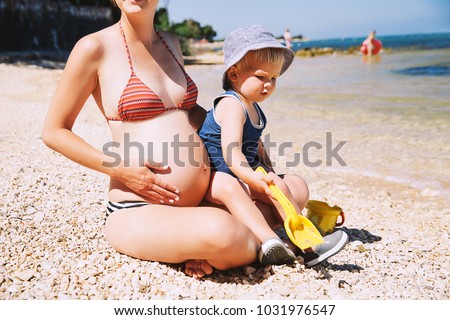 Pregnant mother and her son little cute toddler child playing and relaxing on the sea beach at summer time. Pregnancy, family vacation, parenthood concepts.