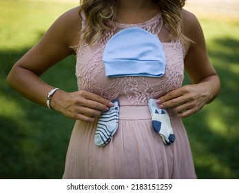 Pregnant Mother Holding A Cap And Two Pairs Of Small Baby Socks On Stomach - Pregnancy Concept.