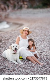 Pregnant Mother With Little Daughter Are Walking Along The Sand Of The Beach Near The Sea With Golden Retriever. Millennial Woman Holds Dog On Leash, Girl By The Hand. Concept Of Traveling With Pets