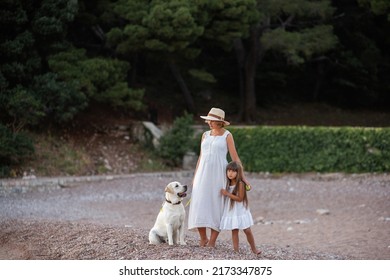 Pregnant Mother With Little Daughter Are Walking Along The Sand Of The Beach Near The Sea With Golden Retriever. Millennial Woman Holds Dog On Leash, Girl By The Hand. Concept Of Traveling With Pets