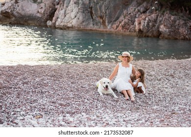 Pregnant Mother With Little Daughter Are Walking Along The Sand Of The Beach Near The Sea With Golden Retriever. Millennial Woman Holds Dog On Leash, Girl By The Hand. Concept Of Traveling With Pets