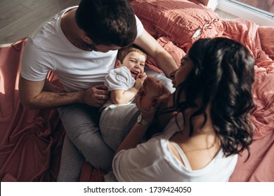 Pregnant Mother Playing With Her Child In The Bedroom. Mother, Father And Their Son Are Sharing Good Emotions While Relaxing At Home Big Window, Natural Light, Terracotta Bed Linen. Family At Home.