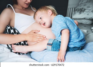 Pregnant mother and son are talking and spending time together in bed at home. Little child boy looking at her mother pregnant tummy. Pregnancy, family, preparation and expectation concepts.