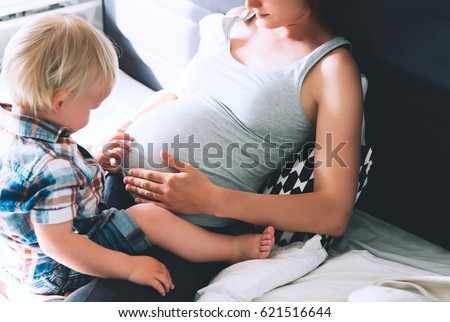 Pregnant mother and son are talking and spending time together at home. Little child boy looking at her mother pregnant tummy. Pregnancy, family, parenthood, preparation and expectation concepts.