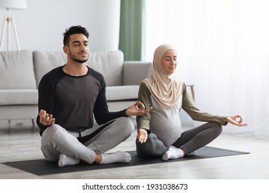 Pregnant muslim couple practicing prenatal yoga at home, meditating together with closed eyes, sitting in lotus pose in living room, enjoying wellness and healthy lifestyle during pregnancy - Powered by Shutterstock