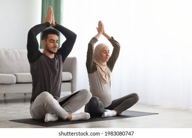 Pregnant Muslim Couple Practicing Yoga At Home, Sitting With Hands Over Head And Closed Eyes On Mat In Living Room, Young Expectant Islamic Spouses Meditating Together, Preparing For Childbirth - Powered by Shutterstock