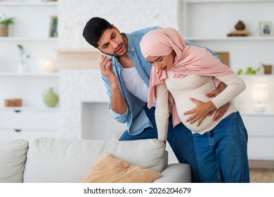 Pregnant Muslim Woman Having Prenatal Contractions, Caring Husband Calling Doctor On Smartphone, Free Space. Young Arab Lady Giving Birth To Baby, Her Partner Arranging Medical Help