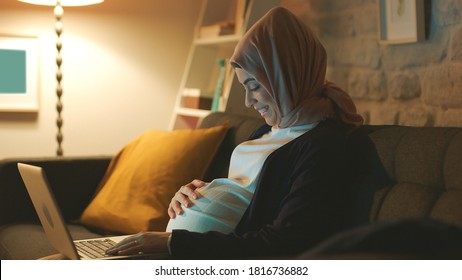 Pregnant Muslim Young Woman Doing Computer Work In Indoor Room With Lights.Concept Of Working At Home.The Woman Loves The Baby She Carries In Her Belly.