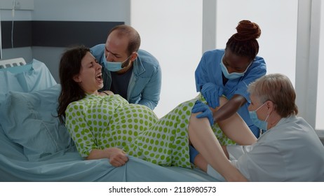 Pregnant Patient In Painful Childbirth Pushing For Baby Laying In Hospital Ward Bed. Multi Ethnic Nurse And Maternity Doctor Giving Assistance To Caucasian Woman For Child Delivery