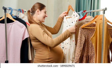 Pregnant Smiling Woman Choosing And Trying Dresses Hanging On Clothes Rack In Her Wardrobe At Bedroom.