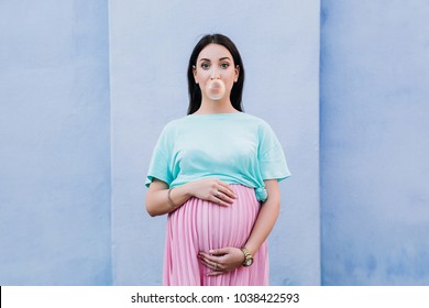 a pregnant stylish girl in a pink skirt and a mint T-shirt hugged her belly with both hands and blows a ball of gum on the background of the blue wall