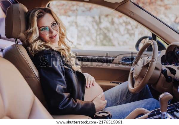 Pregnant swedish woman with wavy blonde hair in round\
glasses, black leather jacket and blue jeans sitting in luxury car\
on drivers sit, touching her belly looking aside with happy smile.\
