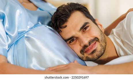 Pregnant wife resting on bed at night and smile as happy that beloved husband care her maternity by tenderly lying down on belly of unborn baby to listen child kicking. Couple enjoy expecting birth.