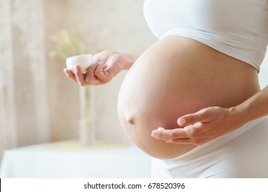 Pregnant woman applying cream at her belly for prevention of stretch marks on the skin in light room