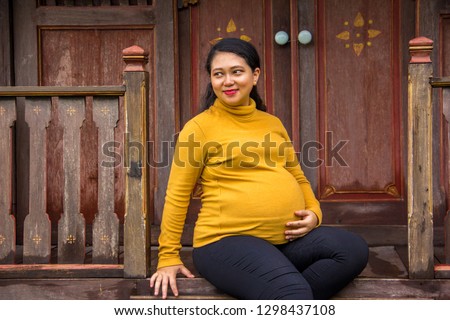 Pregnant woman of Asian ethnicity sits in front of old traditional village house. Side look pose