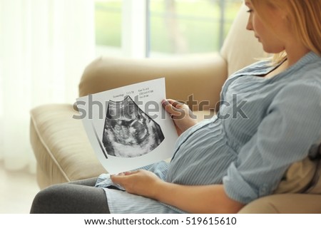 Pregnant woman with baby ultrasound scan sitting on couch at home