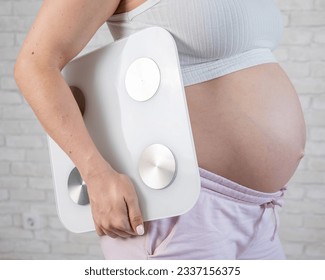 Pregnant woman with bare tummy holding an electronic scale. 