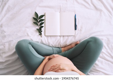 Pregnant woman with beautiful belly makes notes or check list in paper diary. Concepts of preparation for baby birth, tips for a healthy pregnancy. Minimalist style photography. Close-up, indoors.