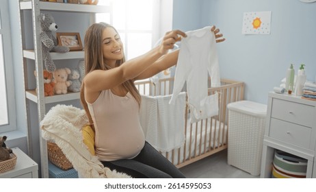 Pregnant woman in bedroom holding baby clothes smiling while sitting near cradle in a cozy home interior with soft toys and natural lighting.
