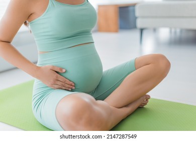 Pregnant woman belly closeup of hands caressing bump wearing maternity activewear during prenatal fitness home class or exercise on yoga mat. Pregnancy lifestyle
