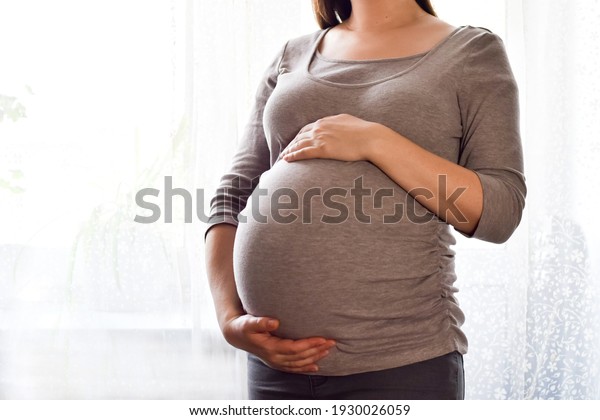 Pregnant woman with big belly at window. Maternity\
prenatal care and woman pregnancy concept. Young pregnant woman\
holds her hands on her swollen belly. Love concept. Horizontal with\
copy space.