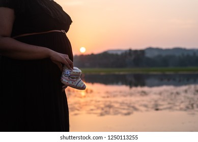 Pregnant Woman In Black Gown Holding Baby Boots And Displays Her Baby Bump At An Outdoor Shoot