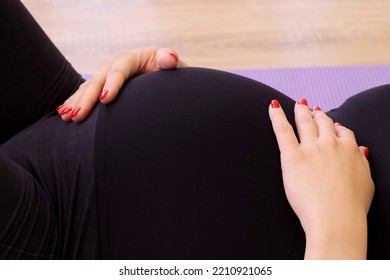 A Pregnant Woman In A Black Tracksuit With Red Nails Lies On Her Back With Her Hands On Her Stomach And Breathes Deeply. Breathing Exercises. Pregnant Yoga Practice Concept