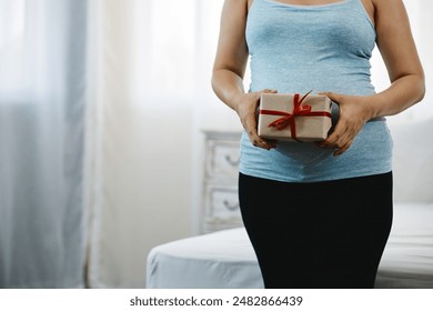 Pregnant woman in blue tank top holding gift box with red ribbon, standing indoors near bed, softly lit room with curtains - Powered by Shutterstock