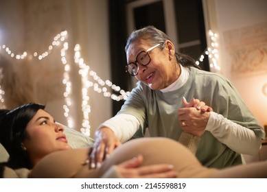 Pregnant woman and caring midwife at home. Woman in casual clothes lying on bed, Asian doula holding hand. Pregnancy, medicine, home birth concept