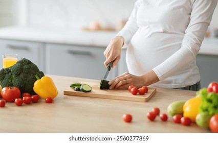 A pregnant woman is chopping cucumbers on a wooden cutting board surrounded by fresh vegetables. The kitchen is bright and inviting, perfect for a nutritious meal preparation, cropped, copy space - Powered by Shutterstock