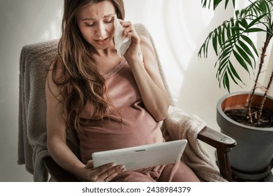  pregnant woman is crying sitting in chair and holding tablet. Rhinitis and increased emotionality in pregnant women watch movie. pregnant woman is crying and wiping her tears with handkerchief - Powered by Shutterstock