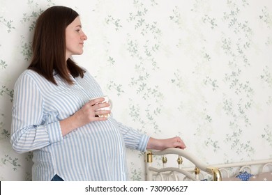 Pregnant woman with a cup of coffee or tea standing near cradle. Anticipation of motherhood concept