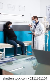 Pregnant Woman Discussing About Childbirth With Specialist During Covid 19 Pandemic. General Practitioner Doing Consultation With Patient Expecting Child At Checkup Visit In Medical Cabinet.