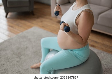 Pregnant woman doing exercises with dumbbells while sitting on a fitness ball at home. Close-up of a pregnant belly. - Powered by Shutterstock