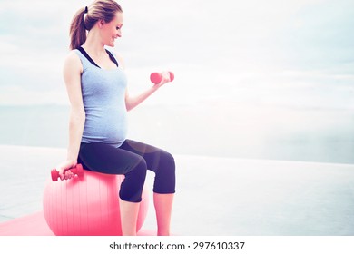 Pregnant woman doing fitness exercises sitting on a pilates gym ball working out with dumbbells, profile view with copyspace - Powered by Shutterstock