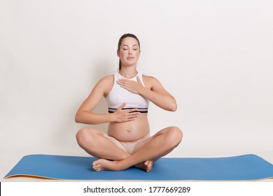 Pregnant Woman Doing Yoga Sitting In Lotus Position Indoors. Future Mother Practice Breathing Exercises With One Hand On Pregnant Belly And Chest, Keeping Eyes Closed.