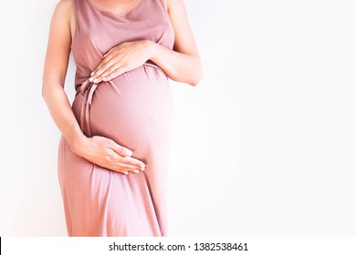 Pregnant woman in dress holds hands on belly on a white background. Pregnancy, maternity, preparation and expectation concept. Close-up, copy space. Beautiful tender mood photo of pregnancy.