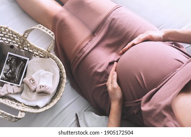 Pregnant woman in dress holds hands on belly. Mother with wicker basket with stuff for newborn, ultrasound image and teddy toy. Expectant mother waiting and preparing for baby birth during pregnancy.