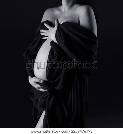 A pregnant woman in a dress holds her belly. A pregnant woman stands on a black background. Pregnant belly. Studio pregnancy photo shoot. Pregnancy. Black and white photo.