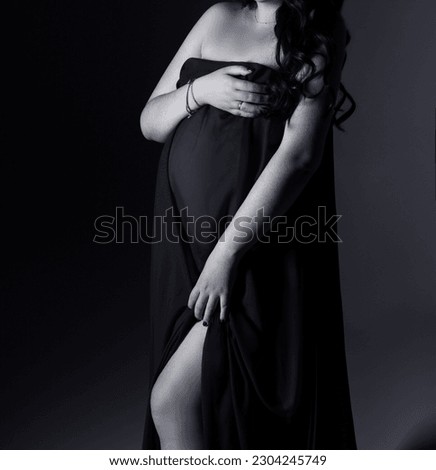 A pregnant woman in a dress stands on a gray background. The belly of a pregnant woman. Studio pregnancy photo shoot. Black and white photo.