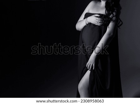 A pregnant woman in a dress stands on a gray background. The belly of a pregnant woman. Studio pregnancy photo shoot. Black and white photo. Beautiful pregnancy.