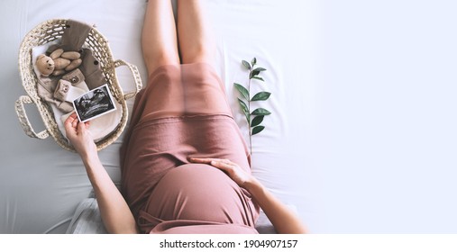 Pregnant woman in dress with ultrasound image. Mother with wicker basket of cute tiny stuff and teddy bear toy for newborn. Expectant mother waiting and preparing for baby birth during pregnancy. - Shutterstock ID 1904907157